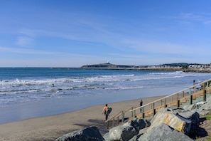 A beach with a wooden staircase leading down to the shore, a rocky outcrop in the foreground, and a distant view of a lighthouse and buildings. - Oceanview Penthouse, Stylish, Walking to Beach (Half Moon Bay)