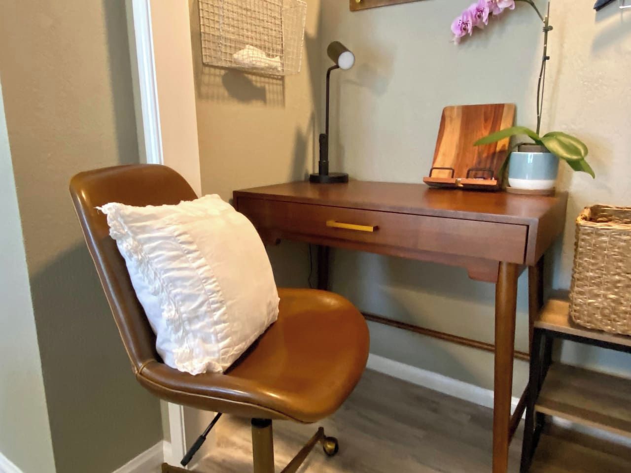 A brown leather chair with a white pillow, a wooden desk with a lamp, a small plant, and a woven basket.