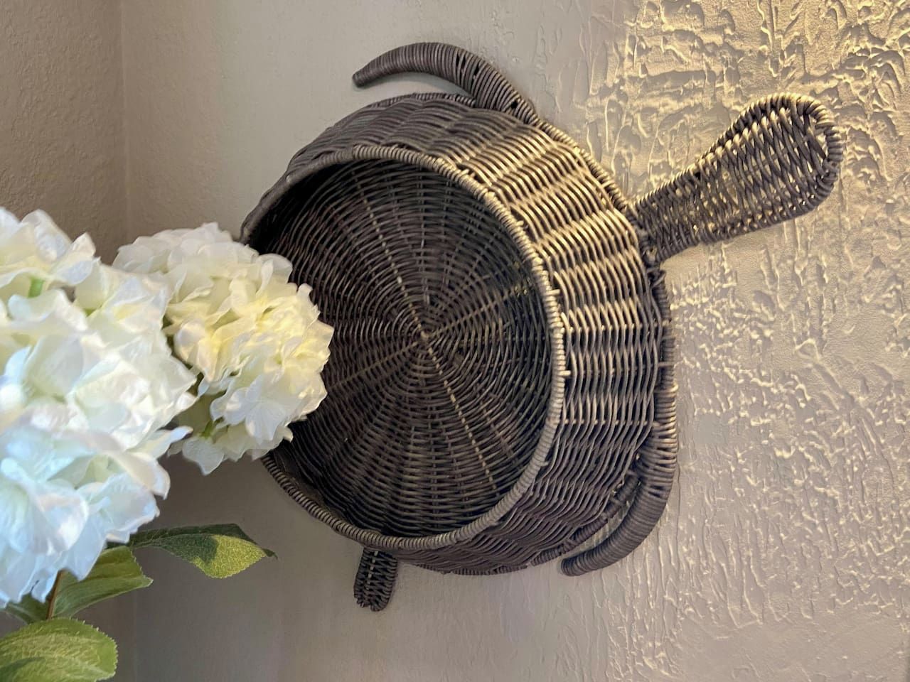 Una cesta tejida colgada en la pared con un arreglo floral blanco a su lado.