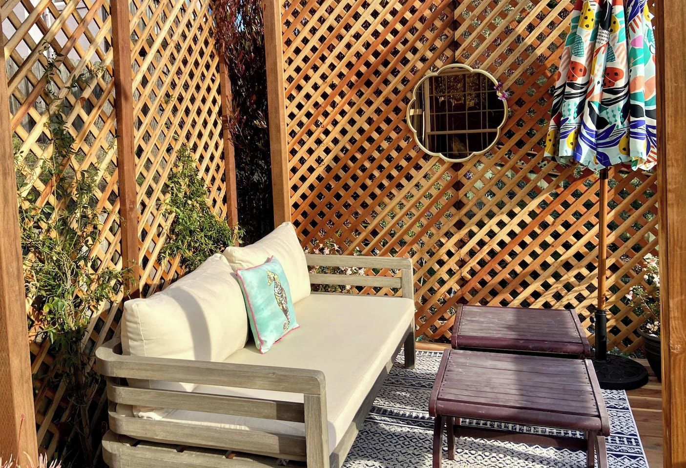A wooden lattice structure with a sofa and a small table, surrounded by plants and a patterned rug.
