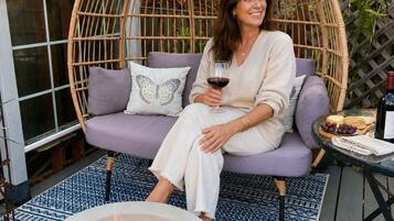 A person seated on a cushioned chair with a glass of red wine, a fire pit in front, and a table with food and drinks to the side.