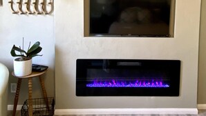 Een woonkamer met een flatscreen-televisie boven een haard met een blauwe vlam. Links staat een bloempot op een klein tafeltje.