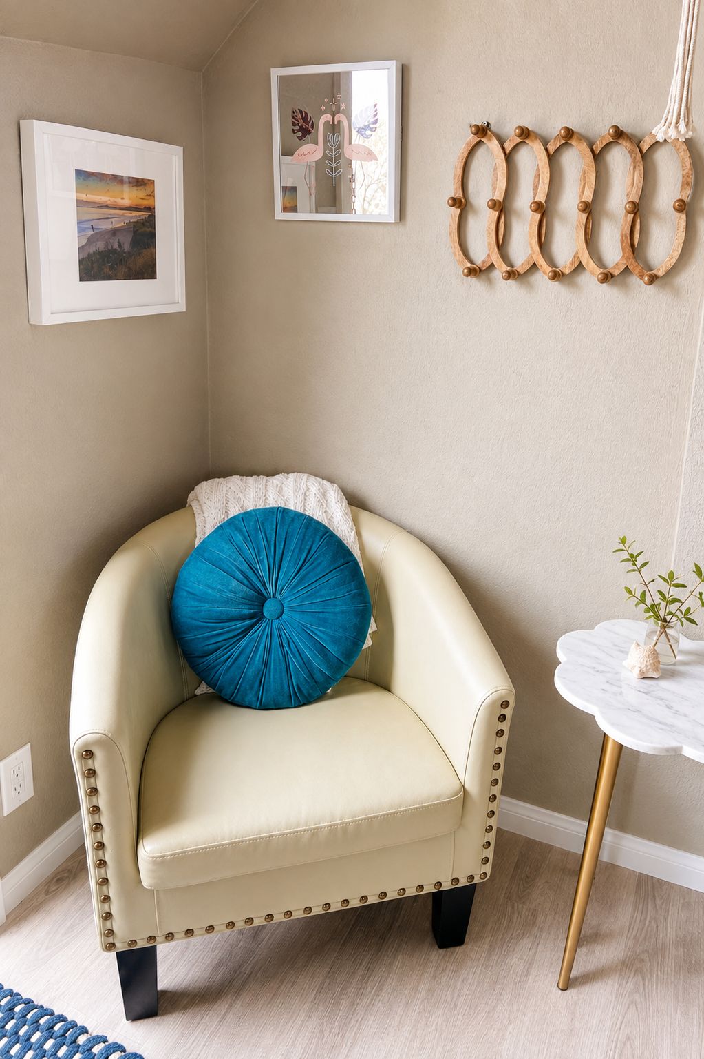 Un fauteuil beige avec un coussin bleu, une petite table blanche ornée d’un vase, et deux tableaux encadrés accrochés au mur.