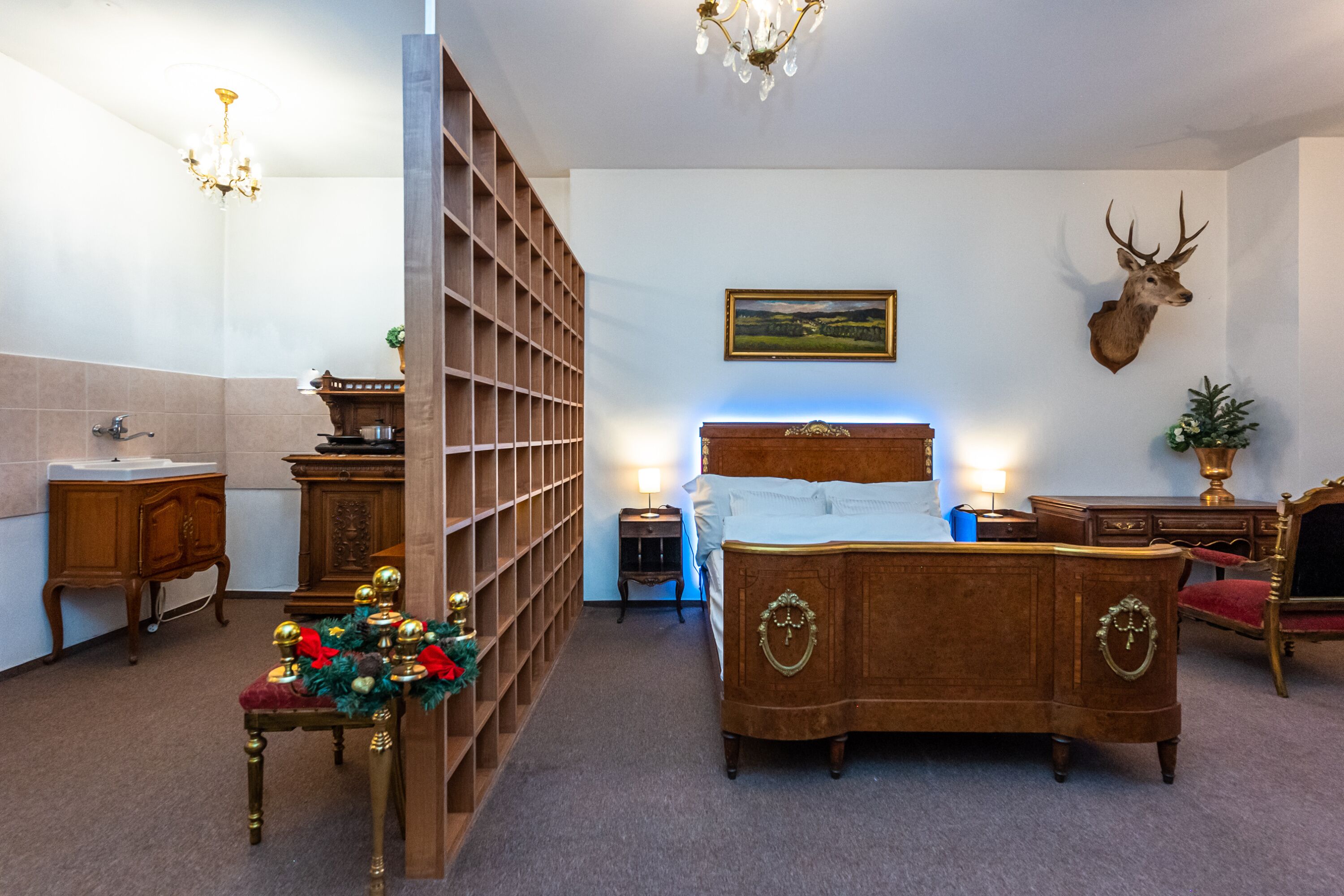 A bedroom with a wooden bed, a mounted deer head, a bookshelf, a chandelier, and a painting on the wall.