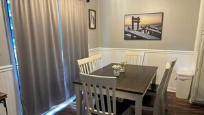 A dining area with a wooden table and white chairs, a framed picture on the wall, and a window with curtains. - Beachfront 2-bedroom condo just steps away from sand, surf and sun. (Myrtle Beach)