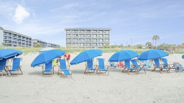 A beach with rows of blue umbrellas and lounge chairs, a large building in the background, and a clear sky. - Beachfront 2-bedroom condo just steps away from sand, surf and sun. (Myrtle Beach)