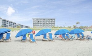 Ein Strand mit Reihen blauer Sonnenschirme und Liegestühle, ein großes Gebäude im Hintergrund und ein klarer Himmel.