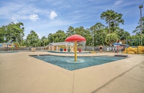 Uma área de recreação com água, com uma fonte central, rodeada por uma piscina e um parquinho cercado.