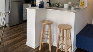 A modern kitchen with white cabinetry, a stainless steel refrigerator, wooden stools, and a blue bench. - Beachfront 2-bedroom condo just steps away from sand, surf and sun. (Myrtle Beach)