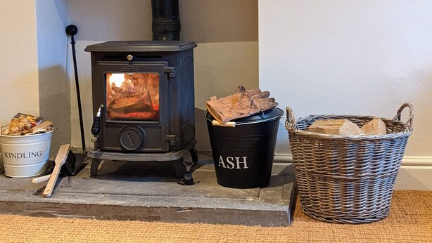 A wood-burning stove with logs, a bucket labeled "ASH," and a wicker basket with firewood.