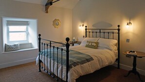 A bedroom with a four-poster bed, a window seat, a small table, and wall-mounted lighting.
