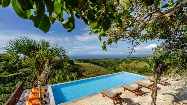 Ein Pool mit Liegestühlen und orangefarbenen Kissen, umgeben von üppiger Vegetation und Blick aufs Meer.