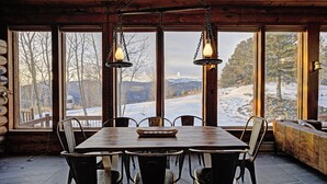 Una mesa de madera para comer con sillas, ventanas amplias que dan a un paisaje nevado y lámparas colgantes.