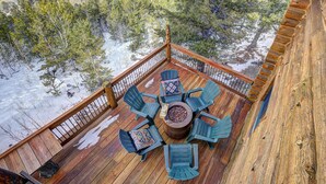 Una terraza de madera con un hoyo para fuego y cuatro sillas, rodeada de suelo cubierto de nieve y árboles.