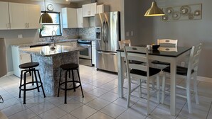 A modern kitchen with white cabinets, a stainless steel refrigerator, a granite countertop, and a dining area with white chairs and a black table.