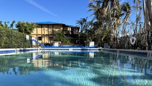 A clear swimming pool with a blue and white tiled edge, surrounded by palm trees and a building with a blue roof.