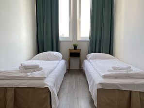 Ein Zimmer mit zwei Einzelbetten, einem kleinen Nachttisch mit Pflanze und einem Fenster mit Vorhängen.