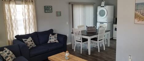 A living room with a dark blue sofa, a wooden coffee table, and a dining area with white chairs and a wooden table.