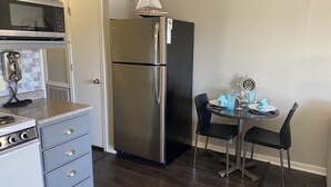 A compact kitchen with a stainless steel refrigerator, a microwave, and a stove. There is a small dining table with chairs and a decorative sailboat on the wall.