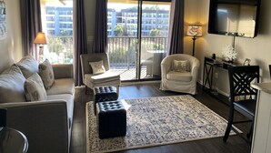 A living room with a sofa, two armchairs, a coffee table, and a rug. There is a balcony with curtains, a ceiling fan, and a TV mounted on the wall.
