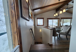 A modern interior with a wooden staircase, a dining area with a table and chairs, and large windows offering a view of the outside.
