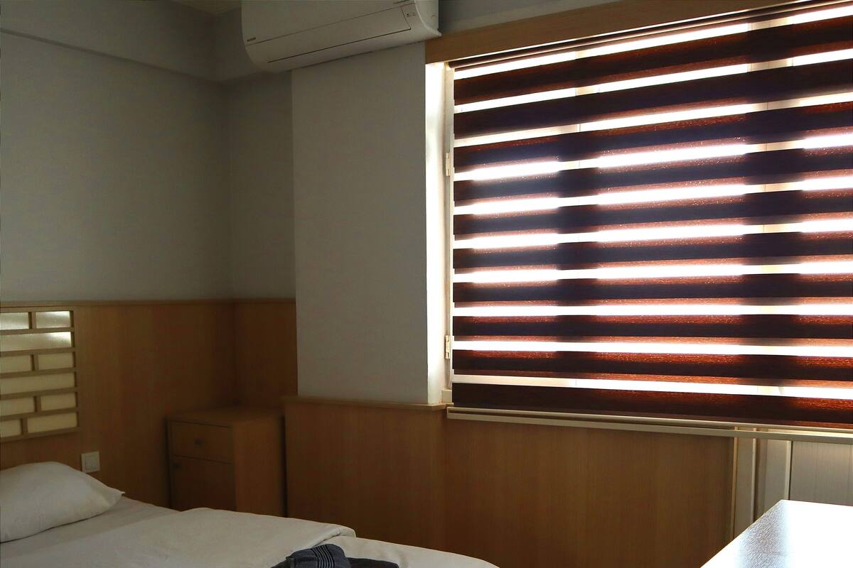 Habitación de hotel con cama, mesita de noche, control remoto y ventana con persianas.