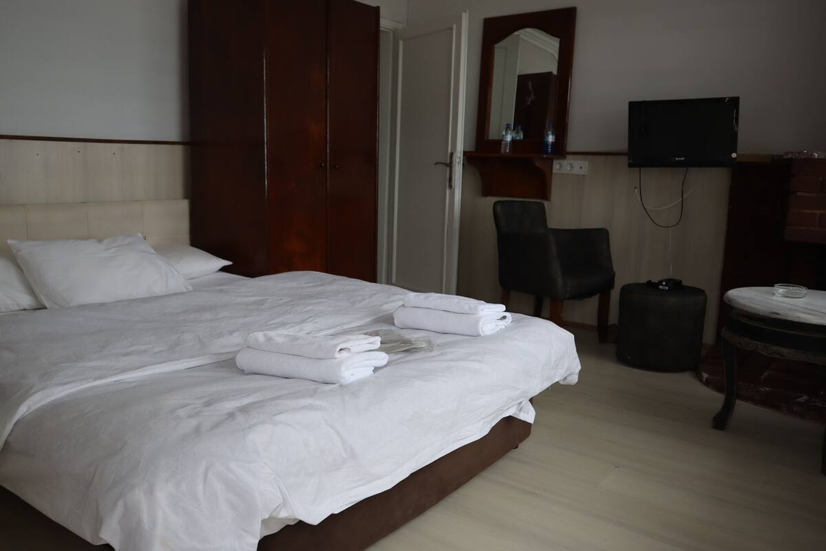 Una cama bien hecha con sábanas y toallas blancas, un armario de madera, una silla, un televisor y una pequeña mesa redonda con un vaso.