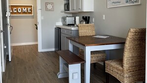 A kitchen with white cabinets, a microwave, and a toaster. There is a wicker chair and a bench at the table. A welcome sign is on the door. - Ocean view Studio, 10 min walk to pier park, right across from Sharkys (Panama City Beach)