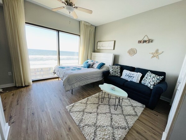 A bedroom with a bed, a nightstand, a dresser, a window with curtains, a ceiling fan, and a view of the ocean.