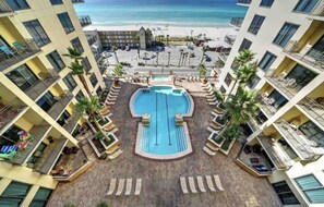 A hotel with a central pool surrounded by lounge chairs, a beach view, and multiple balconies.
