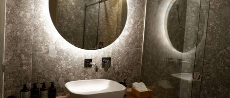 Een moderne badkamer met een groot ovaal wastafel, een ronde spiegel en een douche met glazen wanden.