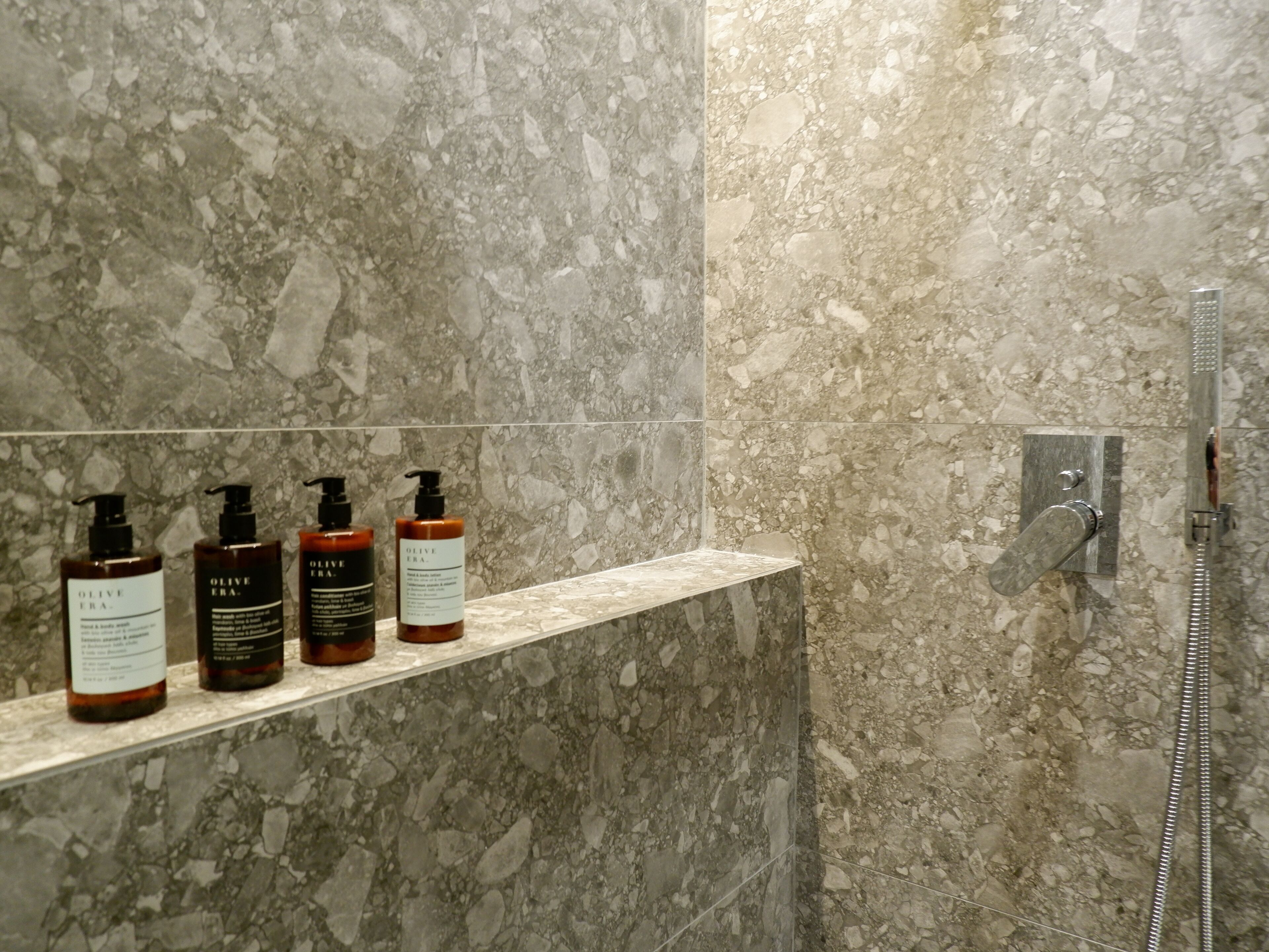 A bathroom with a marble wall, a shelf with three bottles, and a shower control panel.