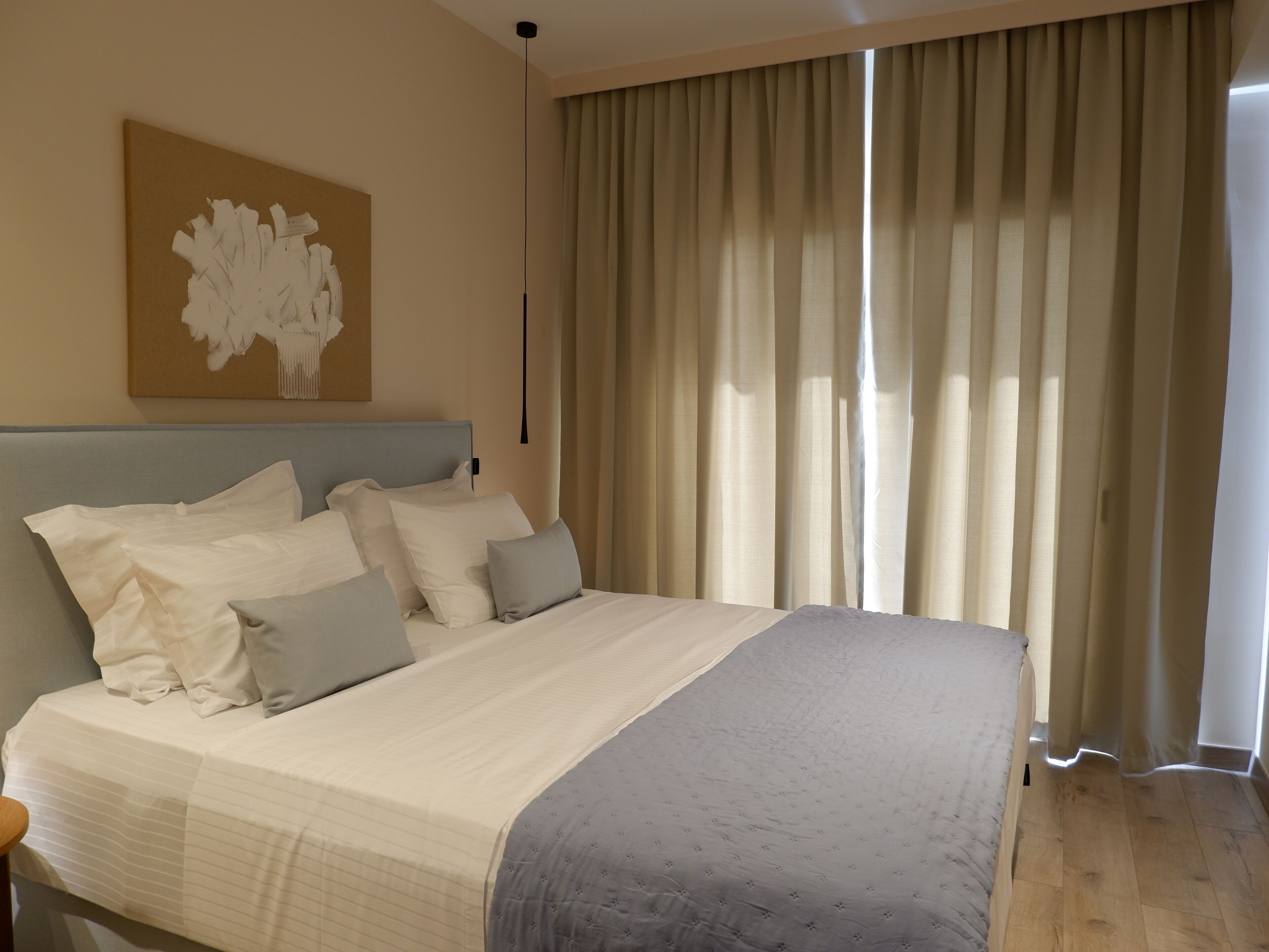 A neatly made bed with a headboard, a large window with curtains, and a wall-mounted artwork.