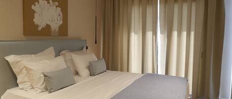 A neatly made bed with a headboard, a large window with curtains, and a wall-mounted artwork.