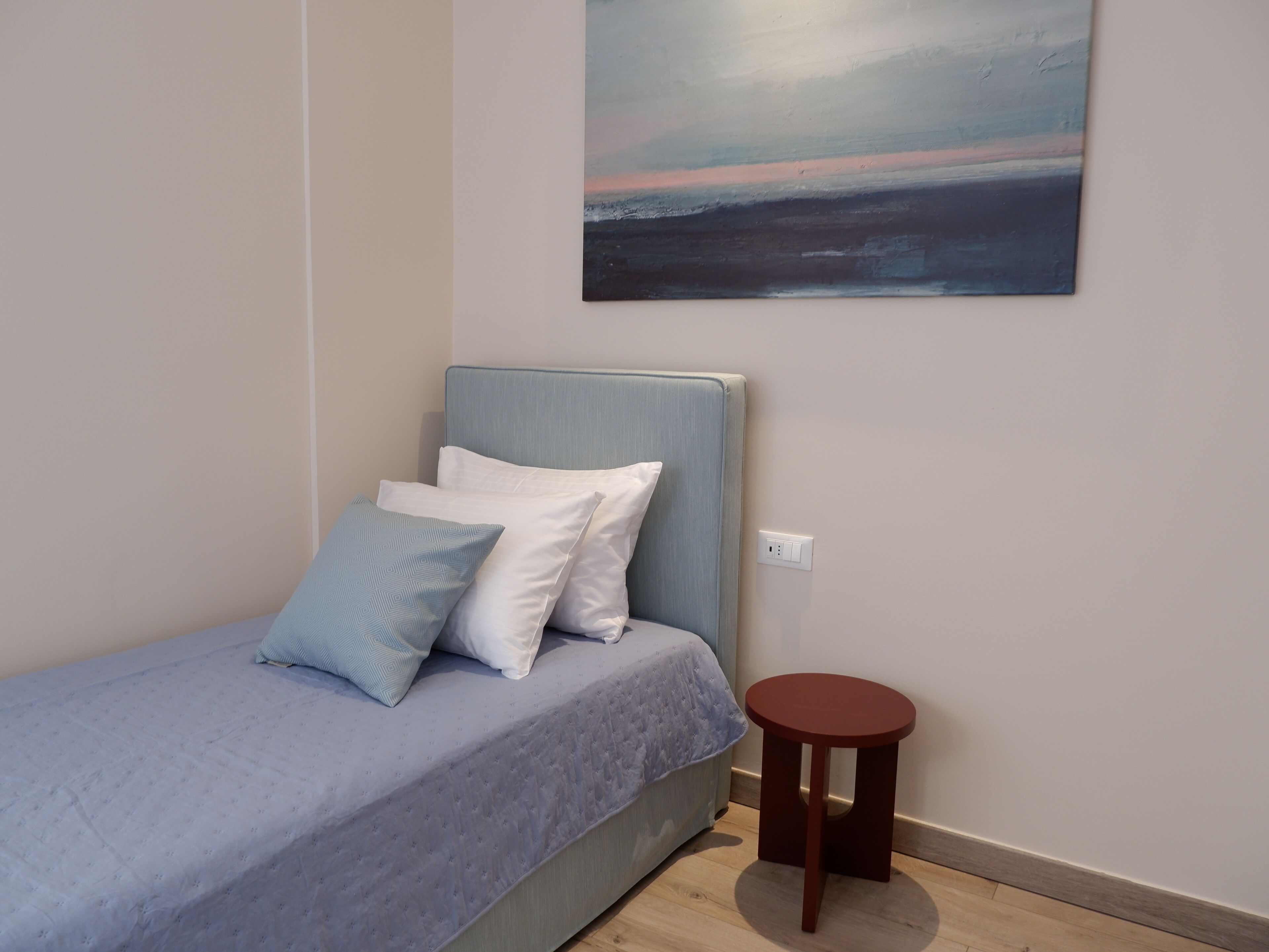 Une chambre à coucher avec un lit, des oreillers, un tabouret rond en bois et un tableau accroché au mur.
