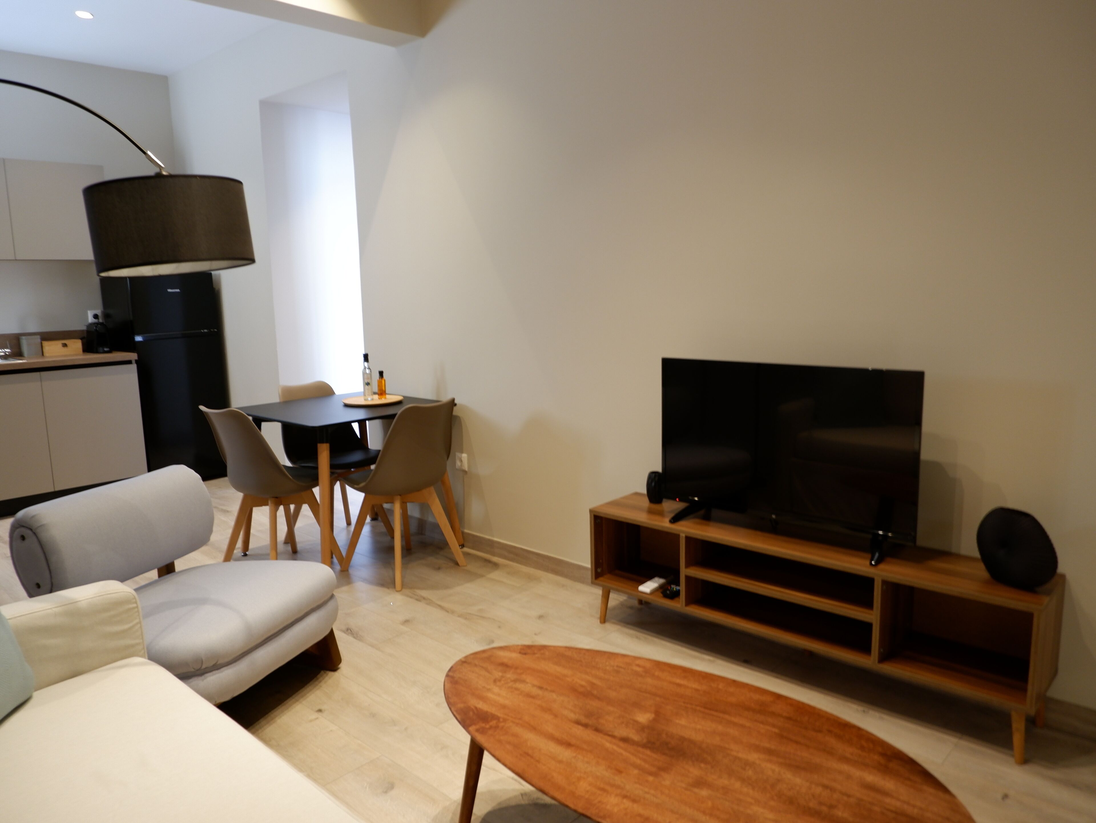 A modern living room with a sofa, a coffee table, a TV on a wooden stand, a dining area with chairs and a table, and a kitchen with a refrigerator.
