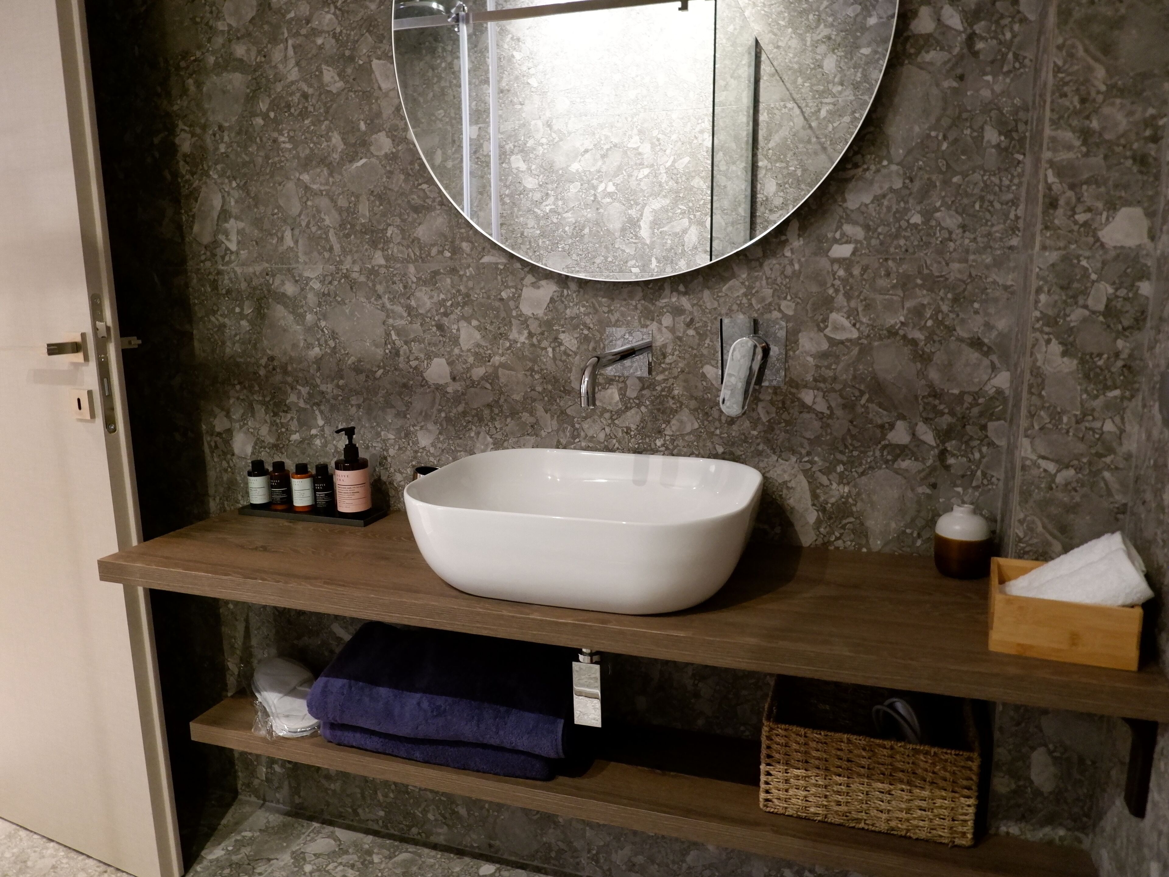 A bathroom with a white vessel sink, a round mirror, and a shelf with toiletries.