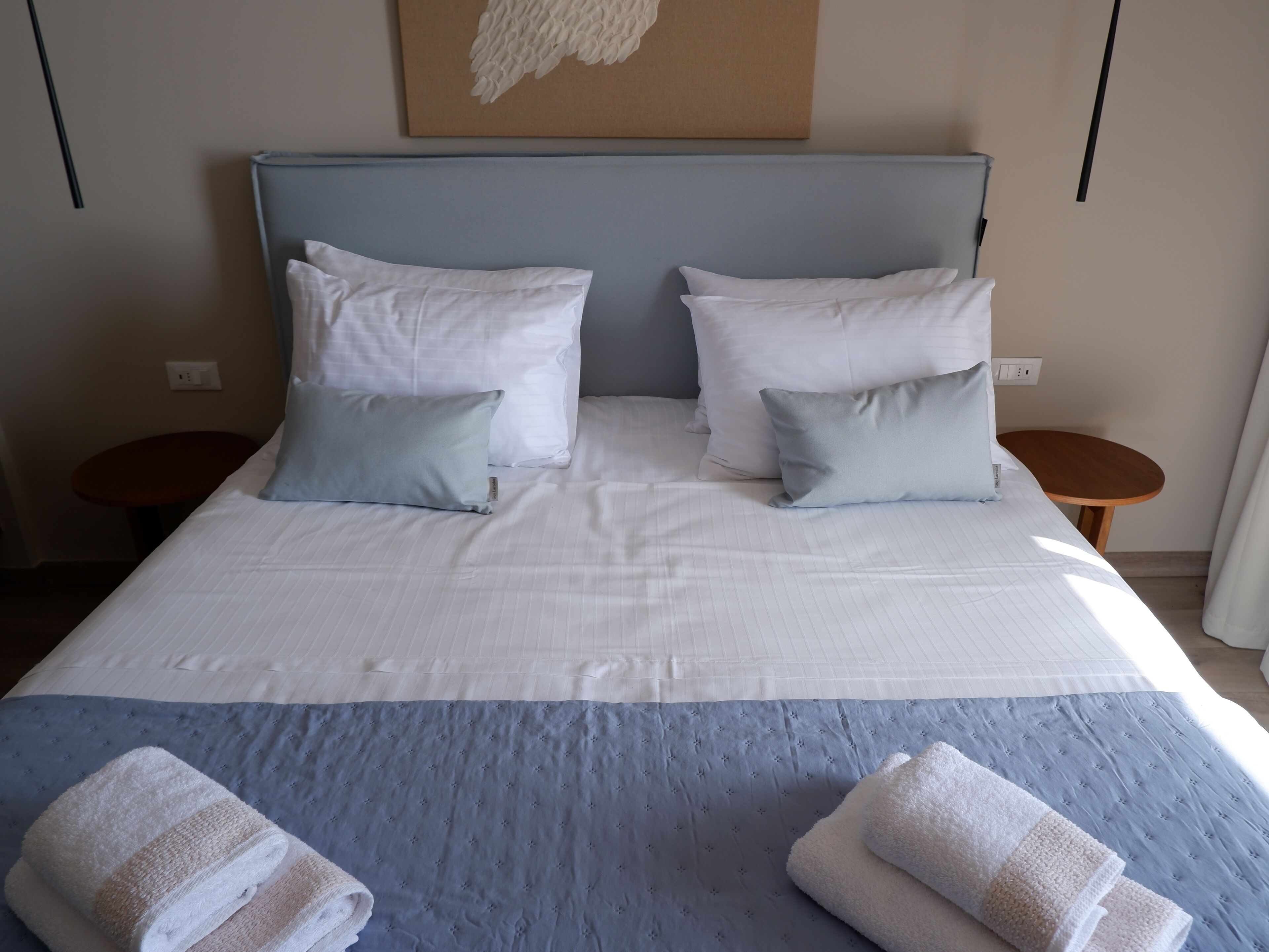 Un lit bien fait, avec une couvre-lit bleue et des oreillers blancs, deux tables de chevet en bois et une œuvre d’art accrochée au mur.