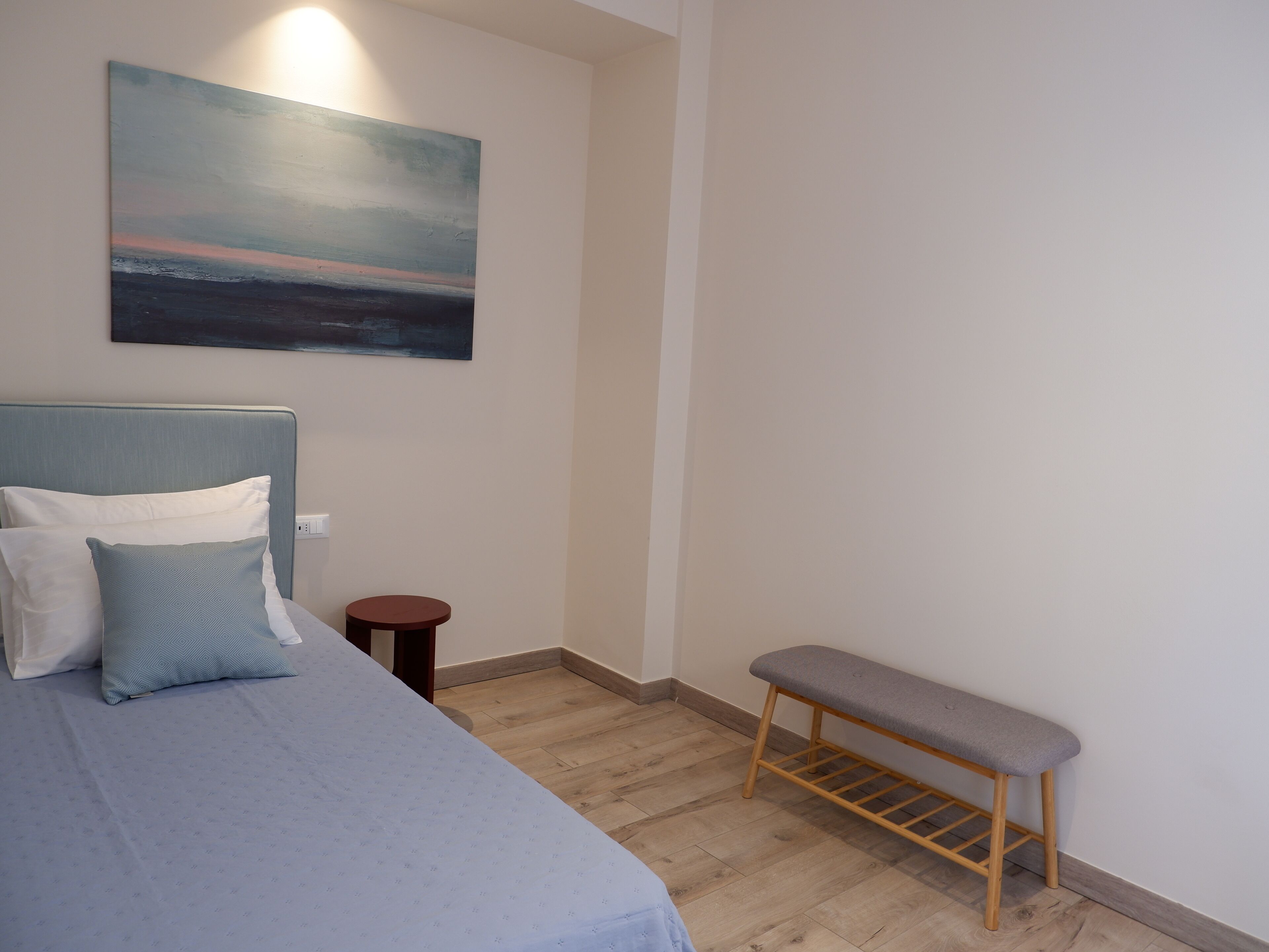Um quarto com cama, almofadas, um banco, uma mesa redonda pequena e um quadro na parede.