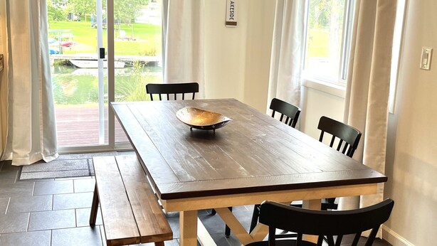 A dining area with a wooden table, black chairs, and a bench. There are curtains on the windows, and a view of a garden outside. - This House Got RIZZ - Recently Renovated Lake Thompson Canal Home (Howell)