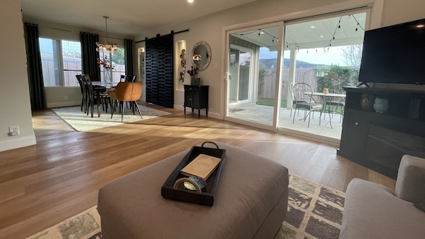 Un salón espacioso con un amplio ventanal corredera que da a un patio, un comedor con mesa y sillas, y un televisor de pantalla plana montado en la pared.
