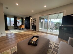 A spacious living room with a large sliding glass door leading to a patio, a dining area with a table and chairs, and a flat-screen TV mounted on the wall. - Lake San Marcos Gem. Walking Distance to Lake, Dining and Golf! (San Marcos)