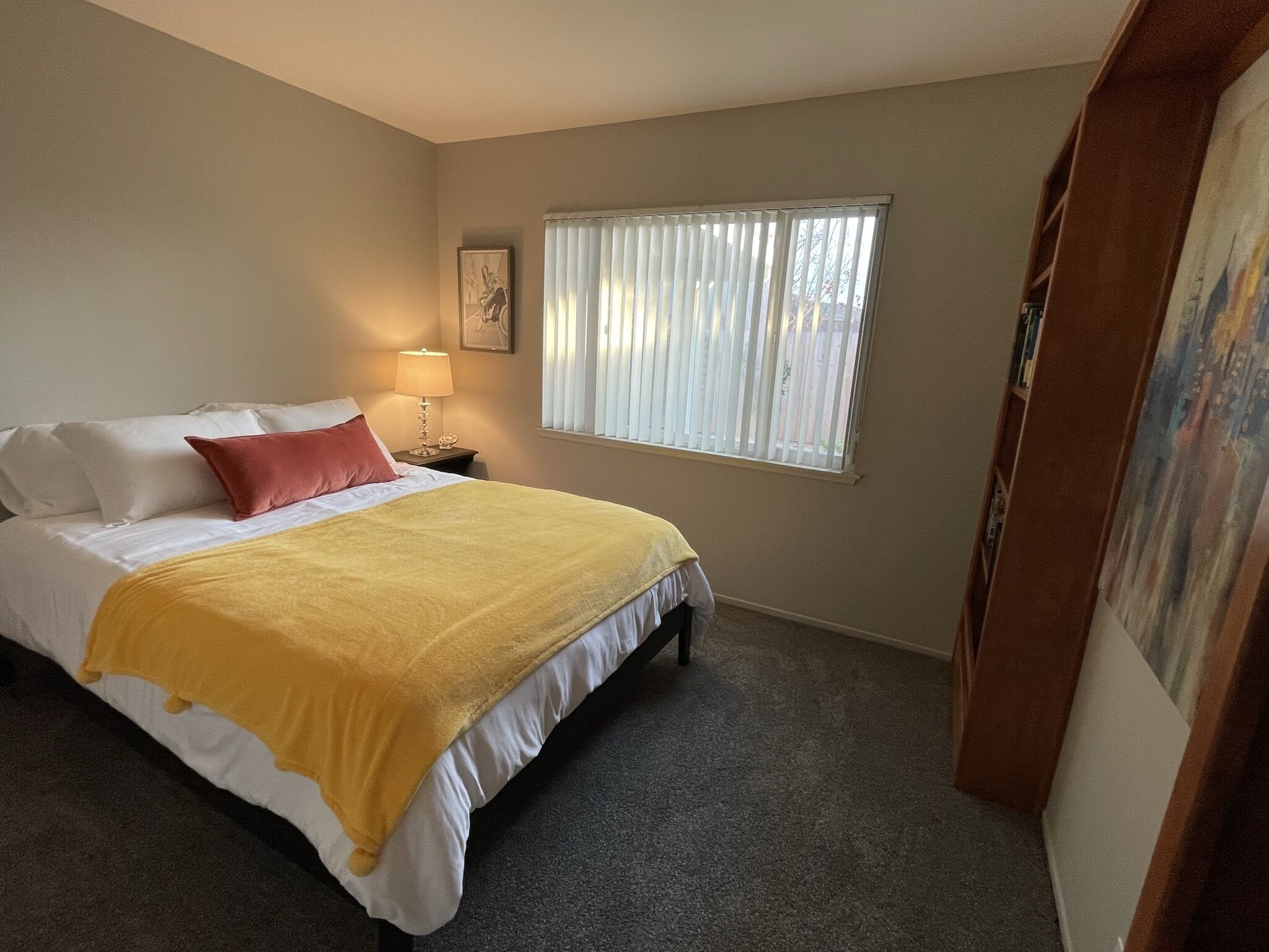 A bedroom with a bed, a nightstand, a lamp, a window with blinds, and a painting on the wall.