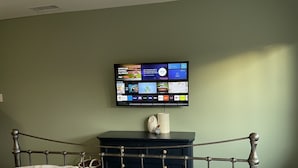Un dormitorio con cama, mesita de noche y televisión de pantalla plana montada en la pared, mostrando la interfaz de un servicio de streaming.
