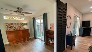 Un salón amplio con suelo de madera, ventilador de techo, un armario de madera, un mueble para televisor y un ventanal grande con cortinas.