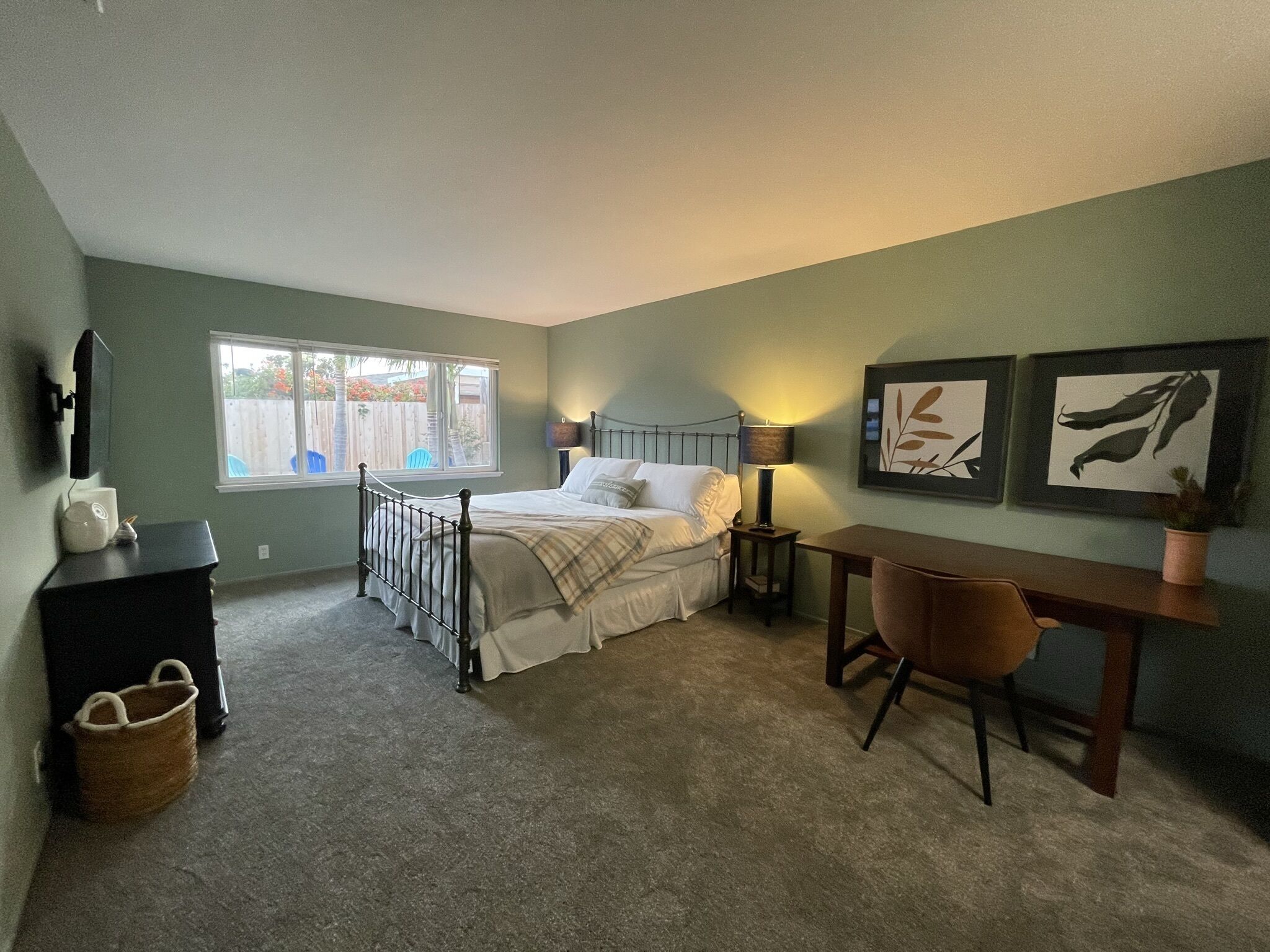 A bedroom with a bed, bedside table, lamp, desk, chair, and artwork on the wall.