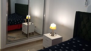 A bedroom with a mirrored closet, a bed with a patterned bedspread, a bedside lamp, and an air conditioning unit on the wall. - APPART CENTRAL LAC 2 T.H.STANDING (Les berges du lac)