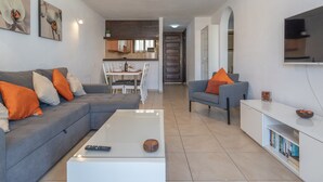 Un salon moderne avec un canapé gris, des coussins orange, une table basse blanche et un téléviseur fixé au mur.