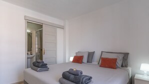 Un lit bien fait, avec du linge de lit gris et deux serviettes enroulées, une table de chevet avec une lampe, et une salle de bain avec un lavabo et un miroir.
