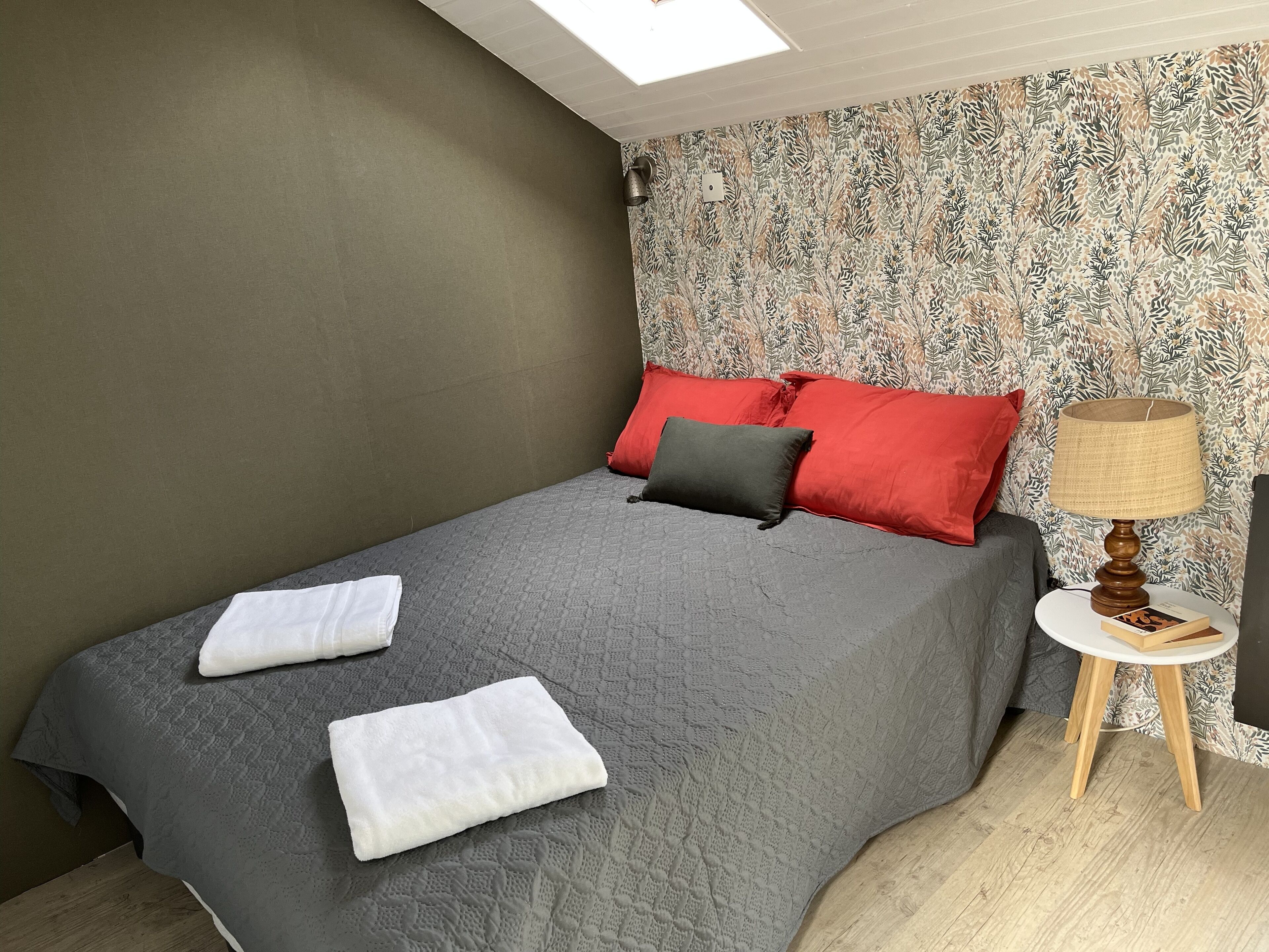 A neatly made bed with grey bedding and red and black pillows, a small bedside table with a lamp, and a patterned wallpaper.